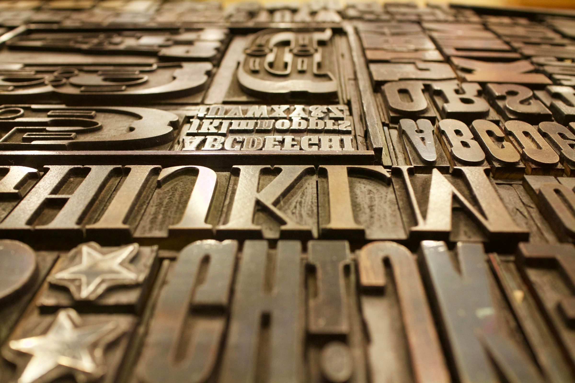 Metal letterpress sorts at a shallow angle, filling the frame: rows of letterforms in relief, the nearest sharp and the rest falling away.